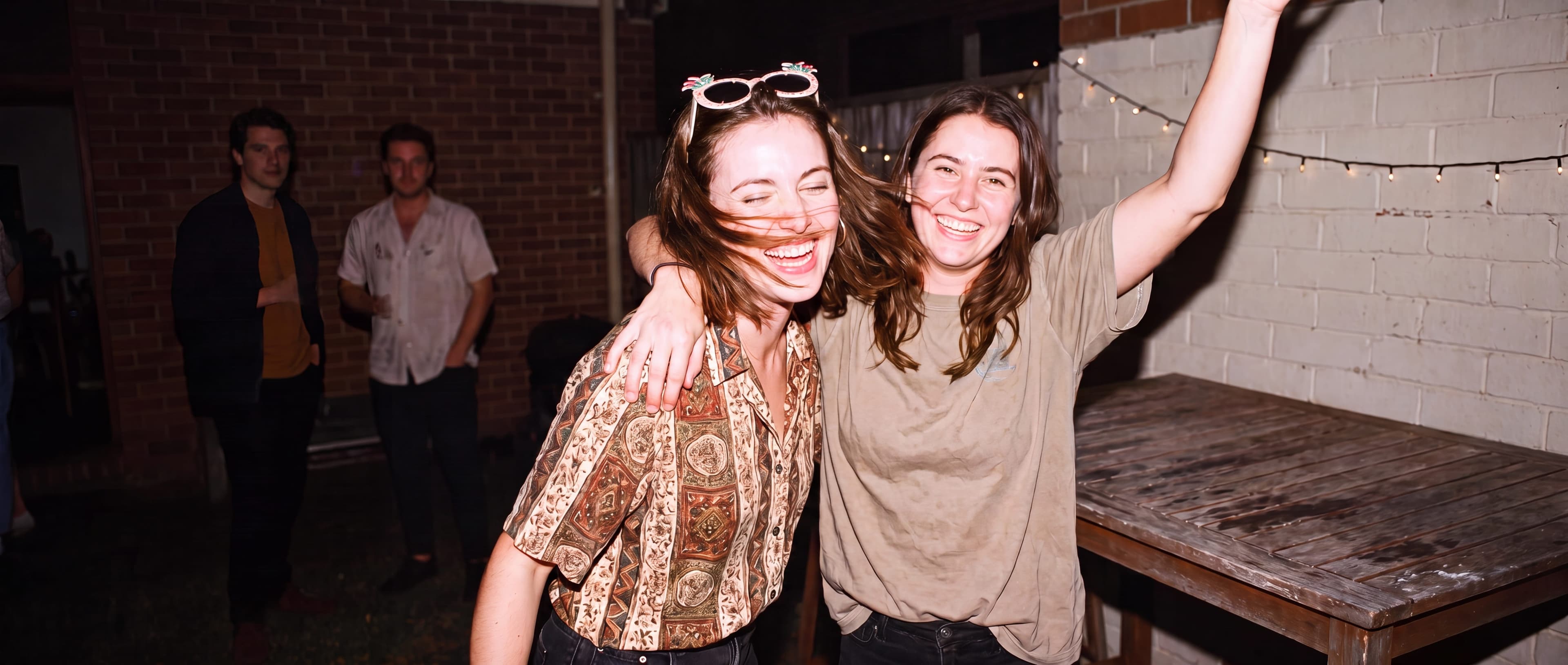 Two friends mid-dance at a small backyard party, one laughing with hair mid-whip, fairy lights on the brick wall behind